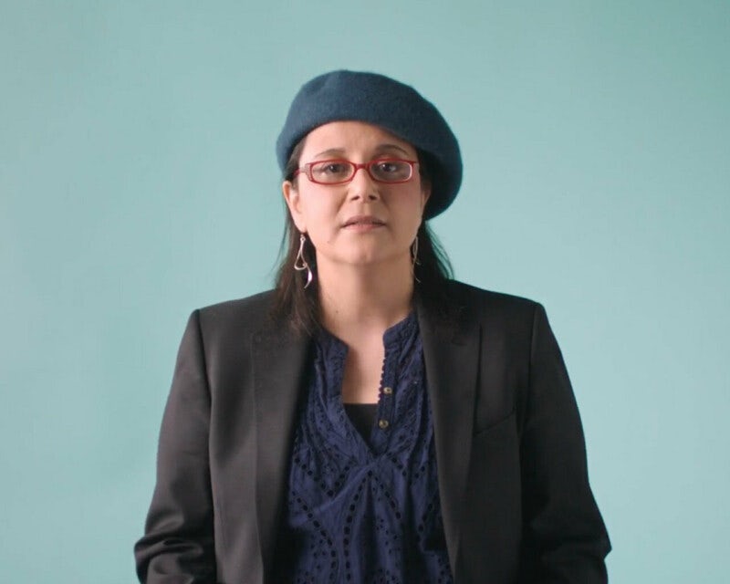 Video still showing a white woman wearing a beret and dark blazer on a teal background.
