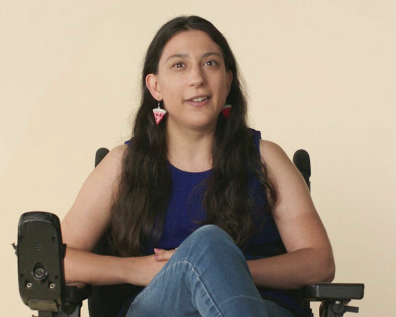 Woman with long dark hair and colourful earrings sits front on to camera on a set. She is sitting in her wheelchair and wearing a dark blue top and blue jeans.