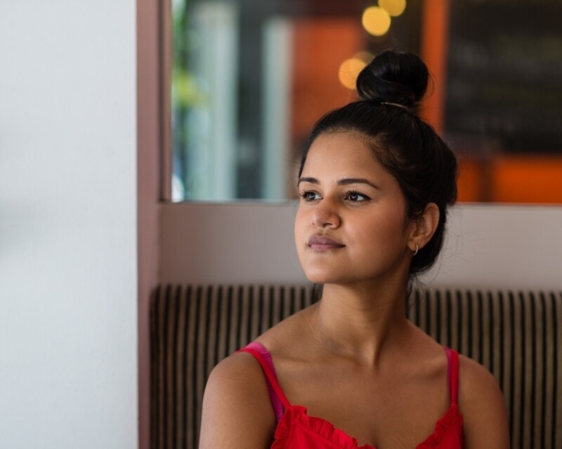 A confident and determined looking young woman with brown skin and dark hair tied up sits in a cafe looking out of a window.