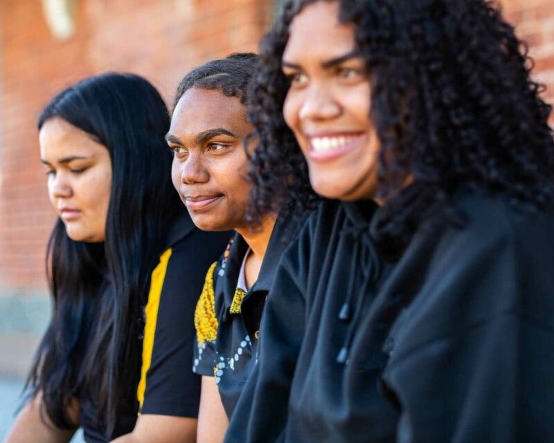 A group of three Aboriginal or Torres Strait Islander women sitting in a row in front of a brick wall, looking away from camera and smiling.