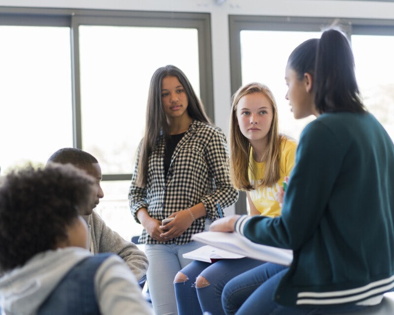 Teenage students sitting on and around a table in a classroom, talking. They are all facing one girl with a pony tail, who is speaking.