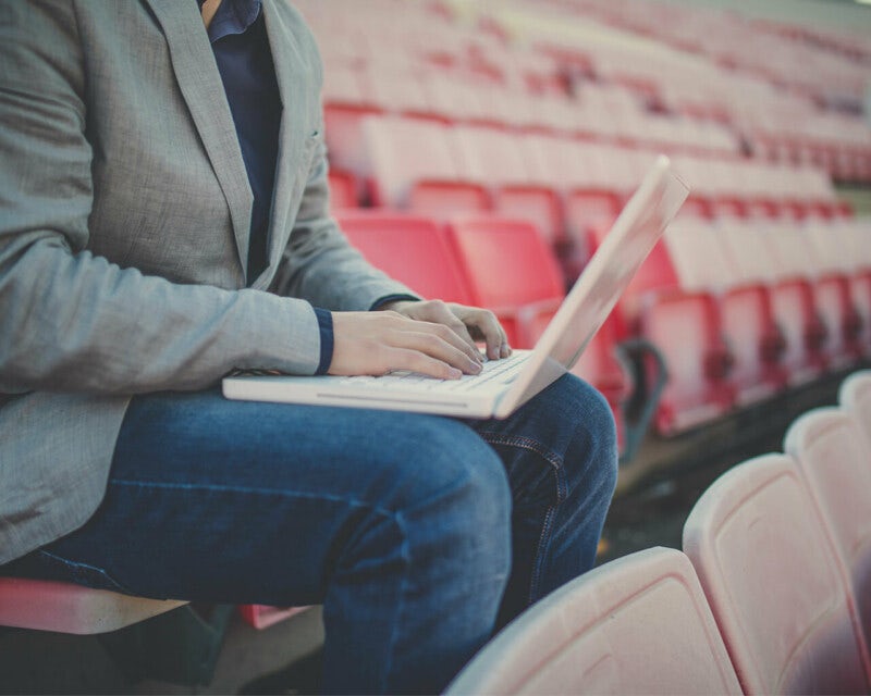 Sports journalists sits on stadium seats using a laptop computer.