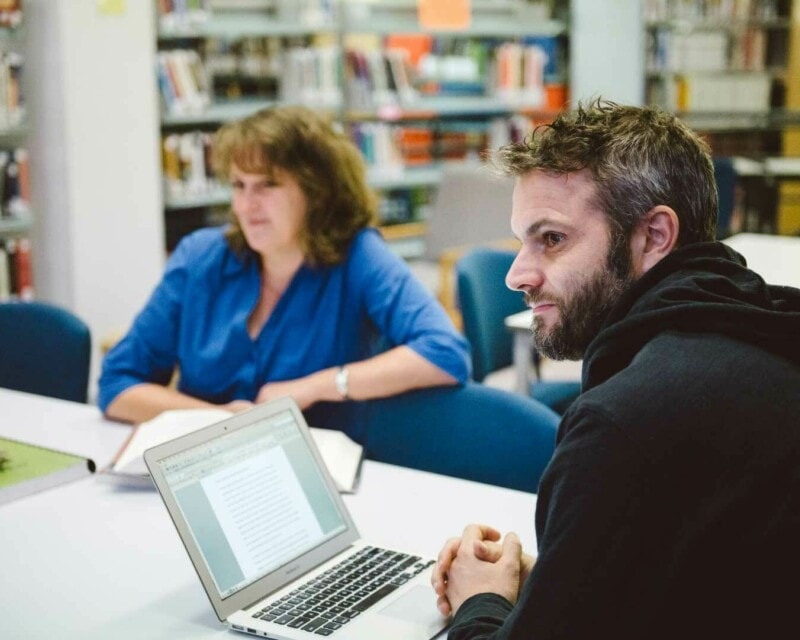 Two mature age TAFE students sitting in a library with laptops and books on the table.