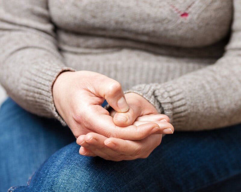 Close up of a woman's hands which she is pressing tightly together. We can just see her jumper, top of her legs and hands.