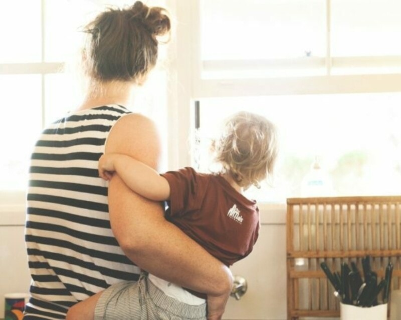 Young woman holding a toddler looking out a window in a bright room. Both their backs are towards the camera.