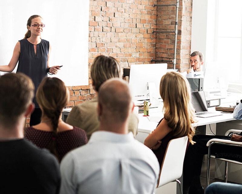 A woman is giving a presentation at a workshop in an office setting to six people. She stands in front of a whiteboard.