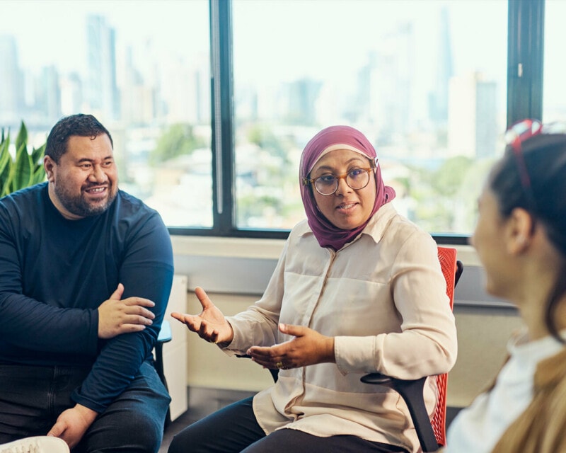 Three people are sitting in an urban office. A woman wearing a headscarf and glasses is speaking to her colleagues who are listening and talking with her.