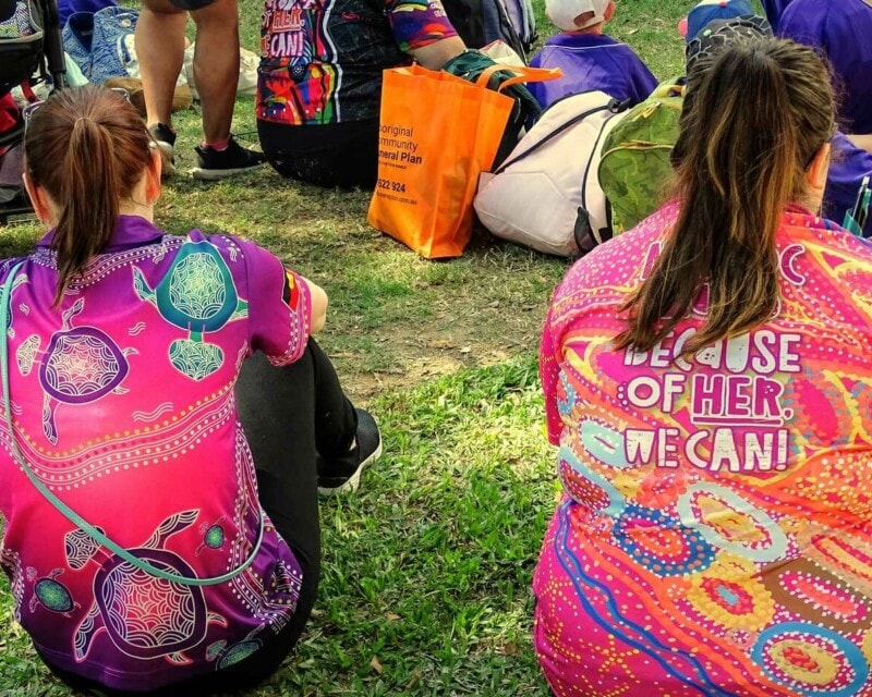 Two women sitting at a festival with their backs to camera. They are both wearing pink tshirts with Aboriginal art designs that say 'Because of her we can'.