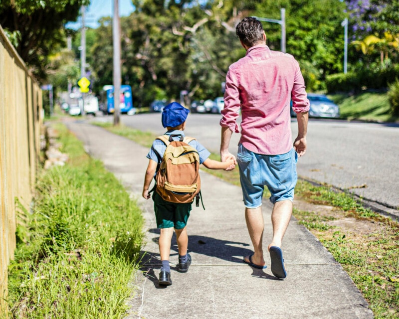 A man in a pink shirt and denim shorts walks his child to school along a suburban street with trees.