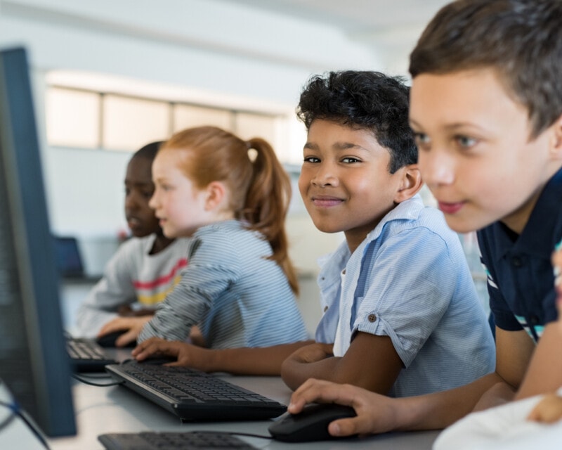 Four primary school aged children sitting at a two computers, sharing keyboards and mice. One child is looking at the camera and smiling.