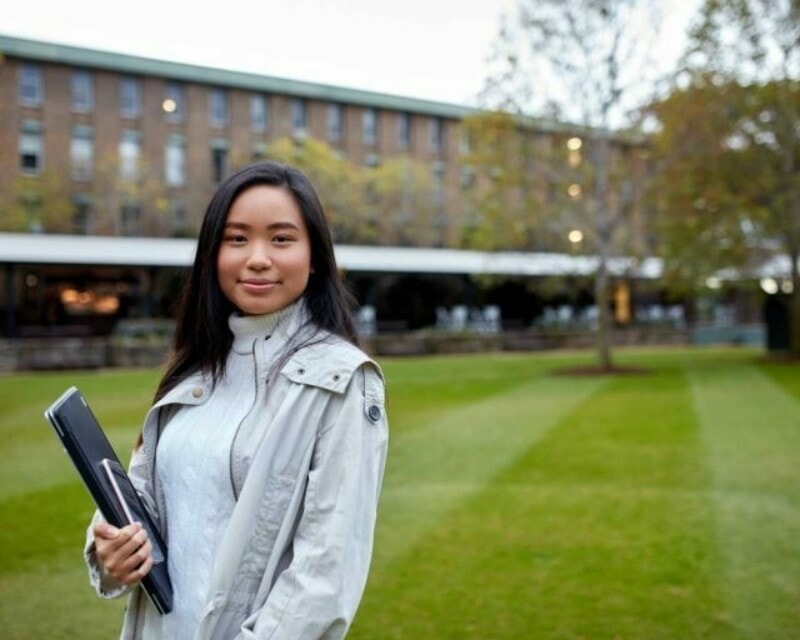 An Asian woman standing on a green lawn at a university campus in front of other buildings. She's holding a laptop and phone.