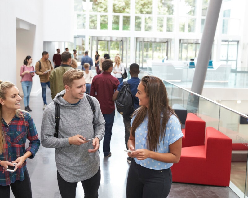 A group of three uni students walk and talk along a light corridor with a high ceiling.