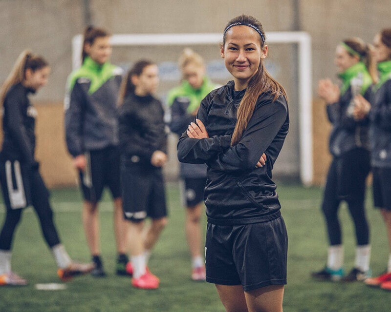 A happy and confident young woman with brown hair stands in front of her soccer team who are chatting. She has her arms crossed over her chest and is smiling.