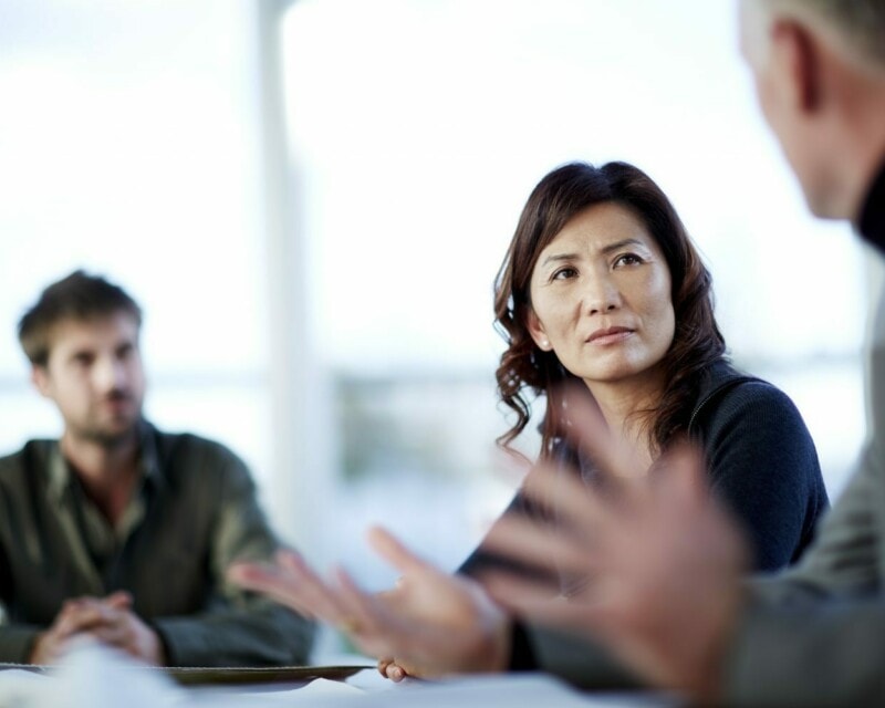 An Asian Australian woman is sitting in a meeting listening to a male colleage, with another male colleage next to her. She is listening intently.