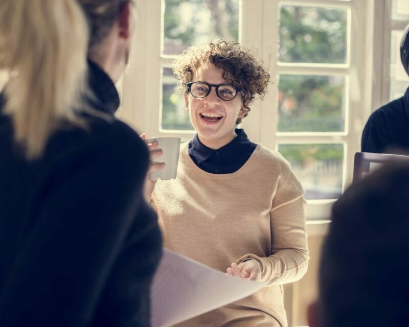 Business people in a casual meeting setting, focus on one woman with curly hair and glasses smiling and drinking coffee.