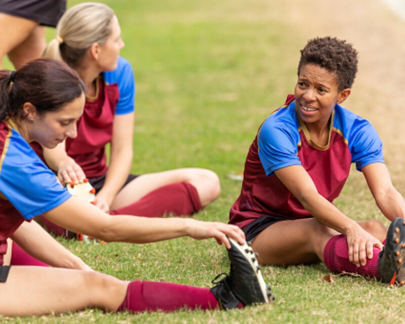 A group of young people in a soccer team sit on the grass to warm up and do stretches before a game.